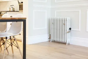 Grace 4 Column cast iron radiator in Ceramic White on feet with nickel valves, against panelled walls beside a kitchen dining table.