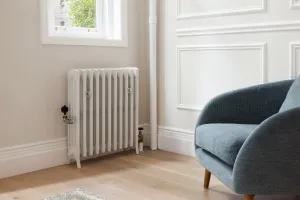 Grace 4 Column 760mm cast iron radiator in Ceramic White on feet under a window, with a black-headed valve, beside a blue armchair on oak floor.