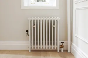 Plain Grace 4 Column cast iron radiator in Ceramic White on feet beneath a window, with brass valves, wall stays and an oak floor.