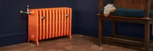 Grace 6 column cast iron radiator in Benjamin Moore Fireball Orange on feet, with a nickel valve, against a navy wall on parquet flooring.