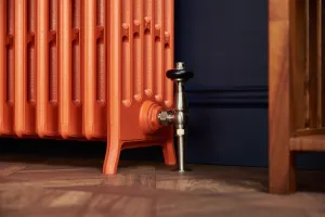 Close-up of a Grace cast iron radiator on feet in Benjamin Moore Fireball Orange 2170-10 with a satin nickel angled valve on a parquet floor.