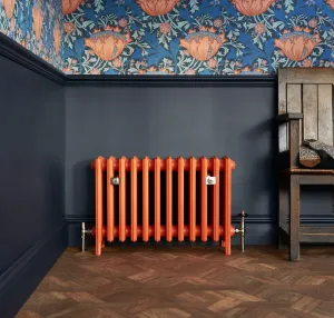 Grace 6 Column 485mm cast iron radiator in Fireball Orange on feet, with nickel valves, below a navy wall, floral wallpaper and parquet floor.