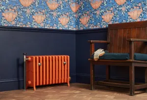 Grace 6 column cast iron radiator in Fireball Orange on feet, with a pipe valve, against navy panelling and floral wallpaper beside a wooden settle.