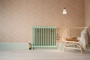 Plain Mercury 2 Column cast iron radiator in pale Pea Green with brass valves, on pink floral wallpaper beside a rattan chair.