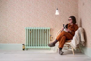Mercury 2 Column radiator in Pea Green 91 with brass valves against pink floral wallpaper, beside a woman holding a dog in a rattan chair.