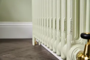 Close-up of a pale green Mercury cast iron radiator with embossed section names, a brass valve with black head, beside white skirting on a wooden floor.