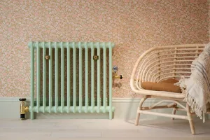 Plain Mercury 2 Column cast iron radiator in Little Greene Pea Green 91 with brass valves, against floral pink wallpaper beside a rattan chair.
