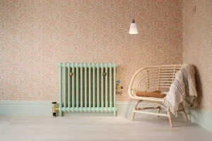 Plain Mercury 2 Column radiator in Pea Green on feet with brass valves, against pink floral wallpaper beside a rattan chair.