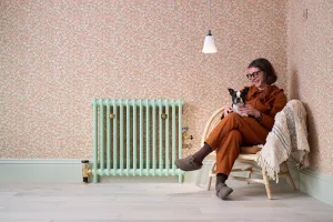 Plain Mercury cast iron radiator in pale Pea Green with brass valves and stays on pink floral wallpaper, beside a woman holding a dog.