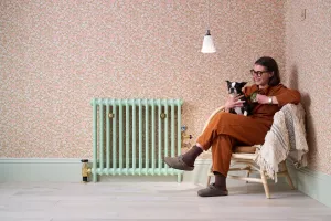Plain Mercury cast iron radiator on feet in Little Greene Pea Green 91 with brass valves, beside floral wallpaper and a woman with a dog.