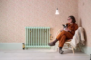 Plain Mercury 2 Column cast iron radiator in Pea Green with brass valves and stays, against pink floral wallpaper beside a woman with a dog.