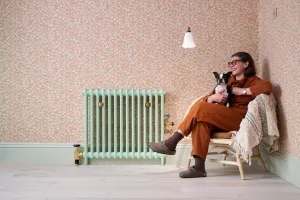 Plain Mercury 2 Column cast iron radiator in Pea Green on feet with brass valves, against floral wallpaper beside a woman holding a dog.