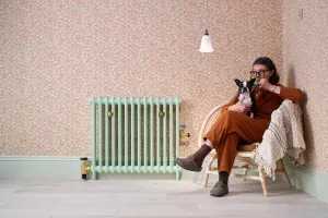 Plain Mercury 2 Column cast iron radiator in Pea Green with brass valves on floral wallpaper, beside a woman with a dog in a rattan chair.