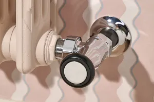 Close-up of a Mercury cast iron radiator in Farrow & Ball Scallop No. 311 with a chrome angled valve and black-rimmed head against wavy pink wallpaper.