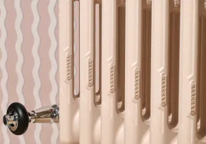 Close-up of a Mercury cast iron radiator in Farrow & Ball Scallop No. 311 with embossed columns and a chrome valve, against pink wavy wallpaper.