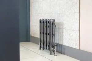 Plain Mercury 3 Column 660mm cast iron radiator on feet in a polished metallic finish, with nickel valves, against a stone-effect wall.
