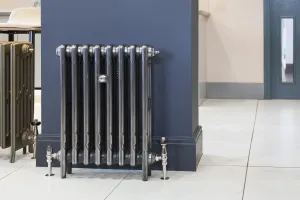 Mercury 3 column cast iron radiator in a burnished metallic finish on feet, with nickel valves, against a navy pillar on a pale tiled floor.