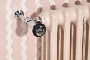 Close-up of a plain Mercury 3 Column cast iron radiator in Farrow & Ball Scallop No. 311 with a chrome angled valve, against wavy pink wallpaper.