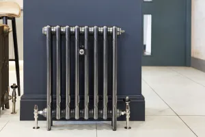 Plain Mercury 3 Column 660mm cast iron radiator in a polished metallic finish on feet, with chrome valves and wall stay, against a navy wall on white tiles.