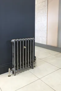 Plain Mercury 3 Column cast iron radiator on feet, polished metallic finish, with nickel valves against a dark blue wall on pale tiles.