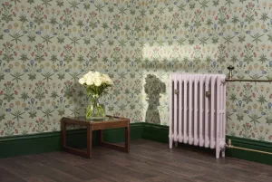 Plain pale pink Mercury 3 column cast iron radiator on feet with natural brass valves, beside floral wallpaper, green skirting and a table of roses.