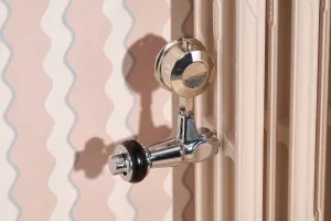 Close-up of a chrome valve with black handwheel on a plain Mercury cast iron radiator in Farrow & Ball Scallop No. 311, against wavy pink wallpaper.