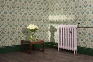 Pale pink Mercury 3 Column cast iron radiator on feet with satin nickel valves, beside a wooden side table of white roses against floral wallpaper.