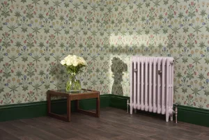 Pale pink Mercury 3 Column cast iron radiator on feet with satin nickel valves, against floral wallpaper beside a table of white roses.