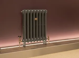 Plain Mercury 3 Column 660mm cast iron radiator in Manhattan on feet, with brass valves, against a plum wall above a lit wooden plinth.