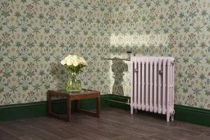 Pale pink Mercury 3 Column radiator on feet with satin nickel valves and wall stays, beside floral wallpaper and a side table with white roses.