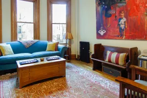 A plain Mercury 3 Column cast iron radiator in Matt Black stands on feet beside a pew bench in a living room with a blue sofa and tall windows.