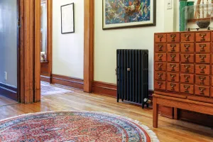 Plain Mercury 3 Column 760mm cast iron radiator in Matt Black on feet in a hallway with oak floor, round rug and wooden filing cabinet.