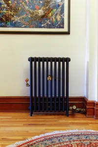 Plain Mercury 3 Column cast iron radiator in Matt Black on feet, with brass valves, on a wooden floor below an abstract painting.