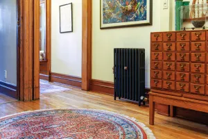 Plain Mercury cast iron radiator in Matt Black on feet against a cream wall, beside a wooden card-index cabinet on an oak floor with a round rug.