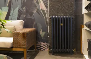 Mercury 3 Column 860mm cast iron radiator in Midnight Ink with brass valves, on feet against a speckled wall beside a wicker sofa.