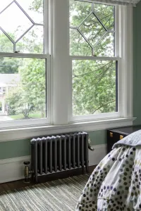 Plain Mercury 4 Column cast iron radiator in Astral Grey Mid with brass valves, on feet beneath a bedroom sash window with green walls.