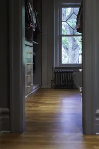Low Mercury 4 Column cast iron radiator in Astral Grey Mid under a sash window, seen through a doorway into a walk-in wardrobe with wooden floor.