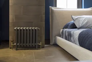 A low plain Mercury cast iron radiator in Astral Grey Mid on feet with brass angled valves, against a tiled wall beside an upholstered bed.