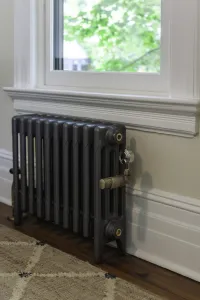 Plain Mercury 4 column cast iron radiator in Astral Grey Mid on feet beneath a white window sill, with brass valves, on a wooden floor.