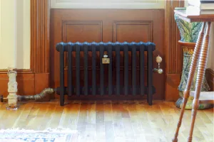 Plain Mercury 4 Column cast iron radiator in Matt Black on feet below a panelled wooden window bay, with brass valves on an oak floor.