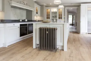 Mercury 4 Column 760mm cast iron radiator in Beaten Brass on feet with angled valves, against a white kitchen island on an oak floor.