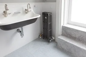 A tall, slim Mercury 4 column cast iron radiator in Astral Grey Mid on feet beside a wall trough sink, white tiles and marble window seat.