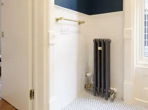 Narrow Mercury 4 Column cast iron radiator in Astral Grey Mid on feet in a white-tiled bathroom corner, with brass valves and a brass towel rail.