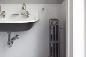 Plain Mercury 4 column cast iron radiator in Astral Grey Mid with a wall stay, beside a wall-hung trough sink in a white-tiled bathroom.