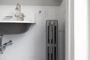 Narrow plain Mercury 4 column cast iron radiator in Astral Grey Mid with a wall stay, beside a wall-hung basin in a white-tiled bathroom.