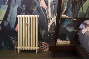 Plain cream Mercury cast iron radiator on feet with chrome valves, against tropical leaf wallpaper beside a bedside table and bed.