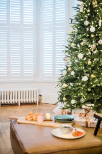 Low white Mercury 6 Column 360mm cast iron radiator with a brass valve, under shuttered bay windows beside a Christmas tree on a herringbone floor.