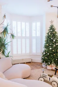 Low white Mercury 6 Column cast iron radiator on feet beneath a shuttered bay window, beside a Christmas tree on a parquet floor.