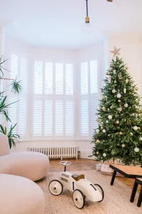 A low white Mercury 6 Column cast iron radiator on feet under a shuttered bay window, beside a decorated Christmas tree and a cream sofa.