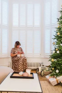 A low white Mercury 6 Column cast iron radiator under a shuttered bay window, a woman seated beside a Christmas tree on herringbone parquet.
